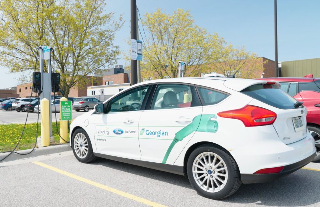 A white electric vehicle that reads "Georgian" on the side is parked in a parking lot in front of an electric vehicle charger. 
