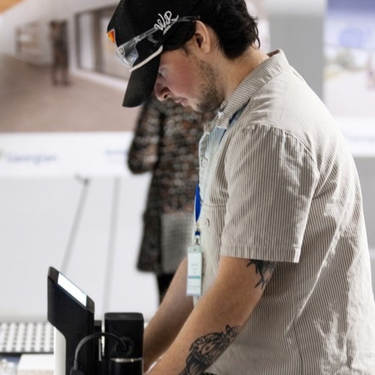 Cabinetmaking Techniques student Matthew Rees works on a piece of equipment in the shop space. He's wearing a black baseball cap and short sleeved beige corduroy shirt. He has dark brown hair and a light beard.