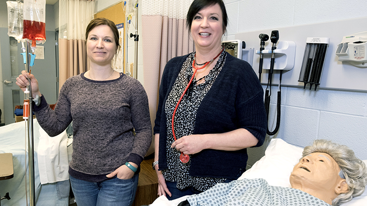 Female-Practical-Nursing-student-and-instructor-smile-at-camera-while-standing-over-a-simulated-dummy-in-the-simulation-lab