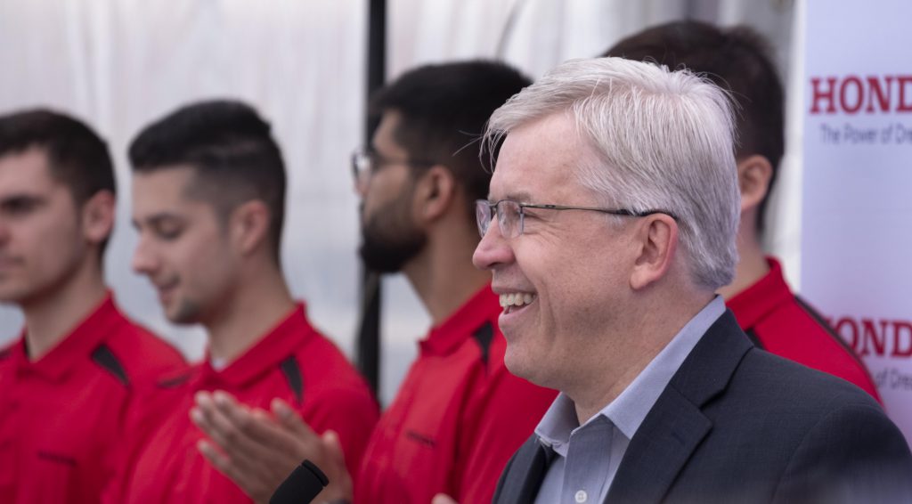 A side angle shot of a gray-haired white man with glasses, wearing a suit. There are young men in red golf shirts behind him.
