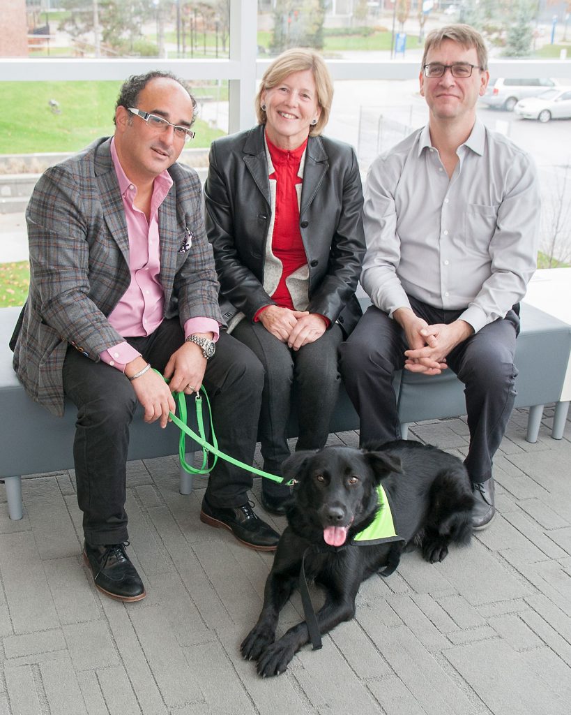 Three people sit on a bench with a black therapy dog on a leash.