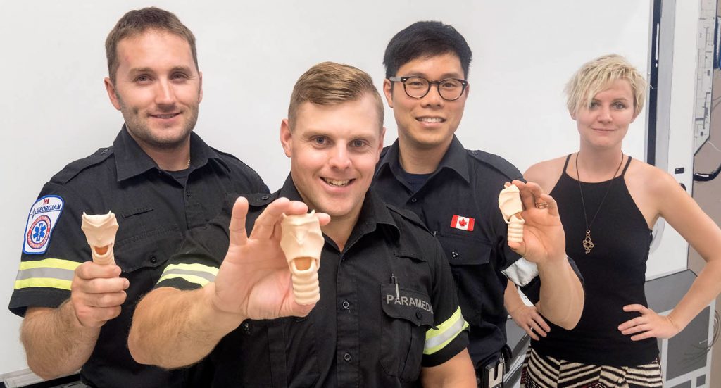 Three young male paramedic students in uniform holding up plastic tracheas and a young blonde woman standing beside them