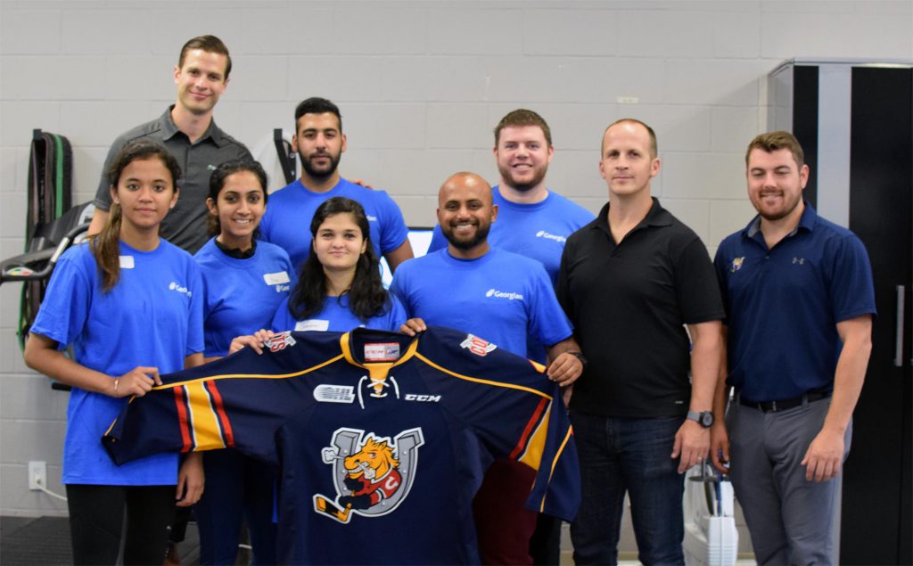 a group of people posing in a classroom. There's some students who are wearing blue Georgian t-shirts and three older men dressed in dark blue golf shirts and pants. The students are holding up a blue hockey jersey that has a horse in a horseshoe logo.