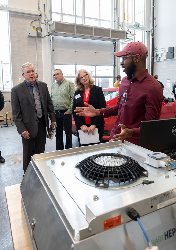 A black male in red clothing demonstrates some equipment to an older man in a suit and a woman in business attire.
