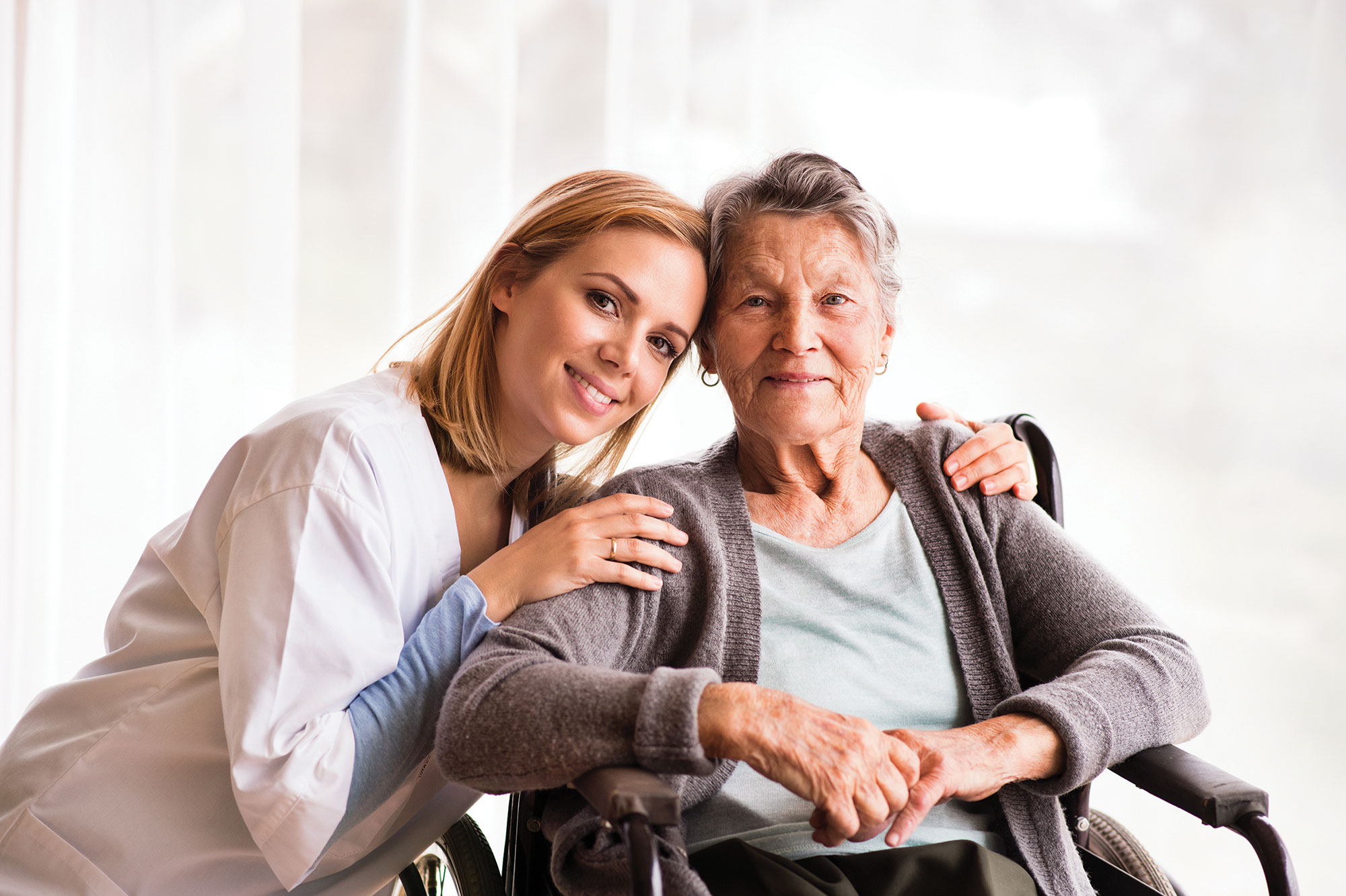 A blonde woman in a white nursing coat kneeling down beside an older woman with short gray hair sitting in a wheelchair. They're both smiling.