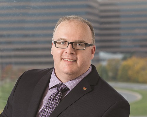 Head shot of a man in black suit with purple tie and shirt, wearing glasses, building behind him