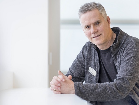 Man in grey sweater and black shirt leans on white counter