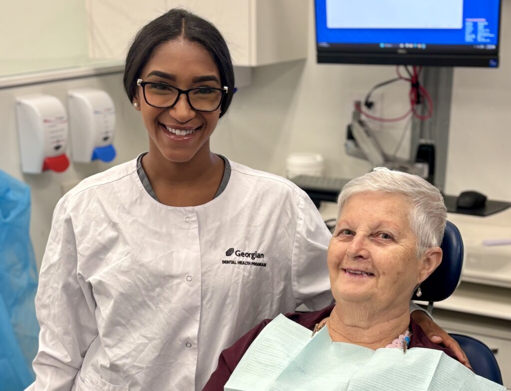 A person wearing a white lab coat reading "Georgian Dental Health Program" smiles next to a person sitting in a dental chair and wearing a blue paper bib.