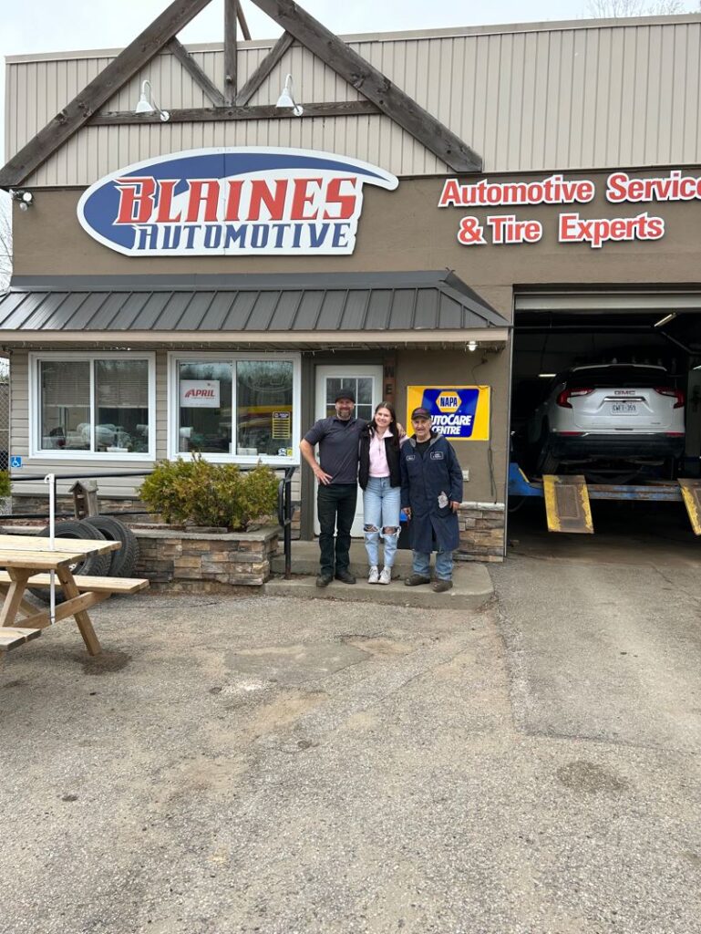 Three people stand out front of an auto shop that has a sign reading "Blaines Automotive."