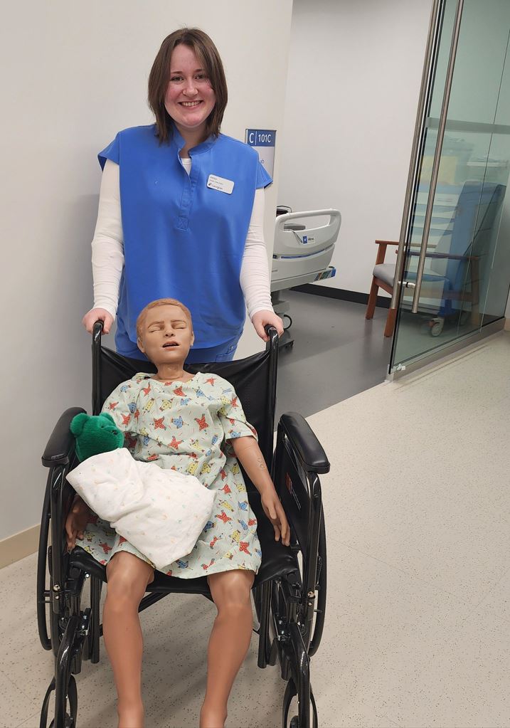 A person wearing nursing scrubs stands behind a wheelchair in which a simulator child patient sits.
