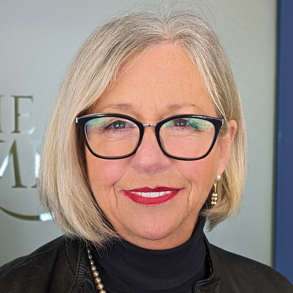 Holly Martin, a 2024 Premier's Awards nominee, smiles at the camera in this headshot. She has light blond/silver hair which is worn in a chin-length bob. She's wearing dark-rimmed eyeglasses, dark pink lipstick and a black turtleneck sweater.