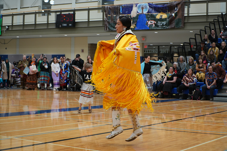 A dancer dressed in a yellow Indigenous regalia dancing in a gym