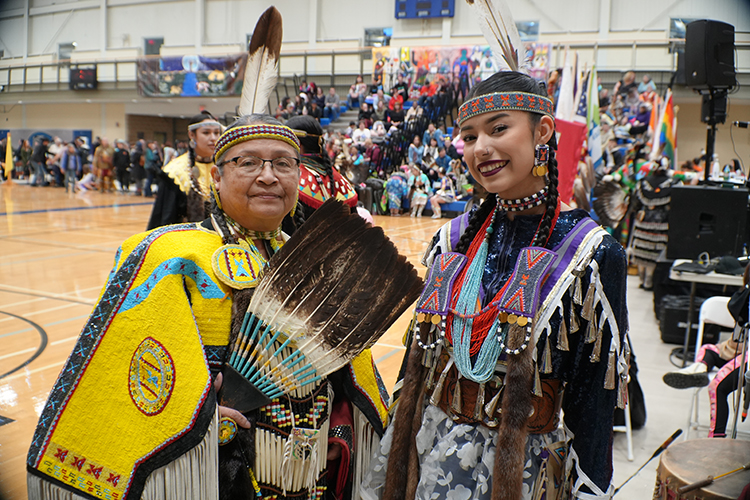 Two females dressed in Indigenous regalia smiling and standing in a gym