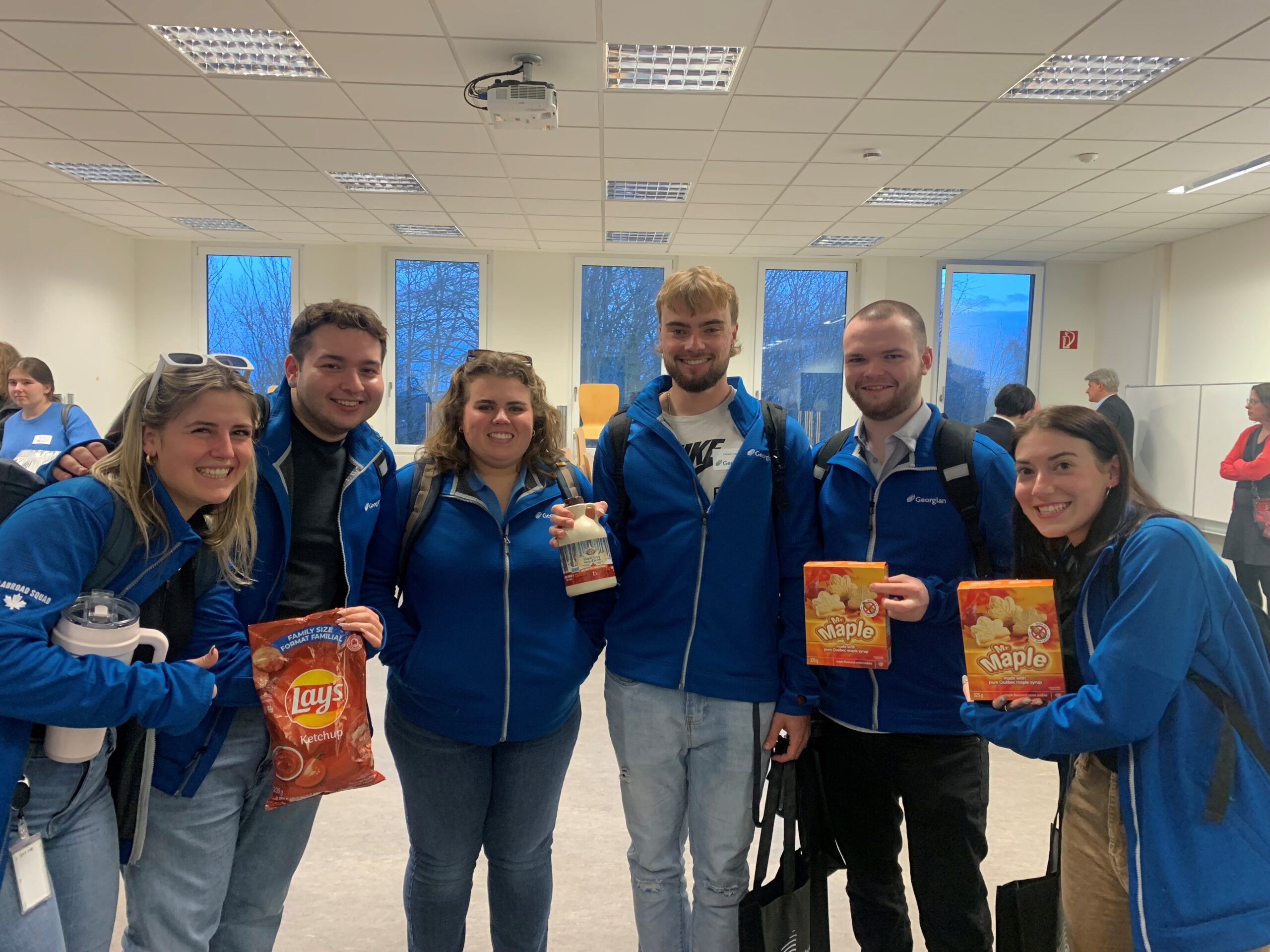 鶹ֱ students in blue 鶹ֱ jackets holding Canadian treats for International Flavours activity.