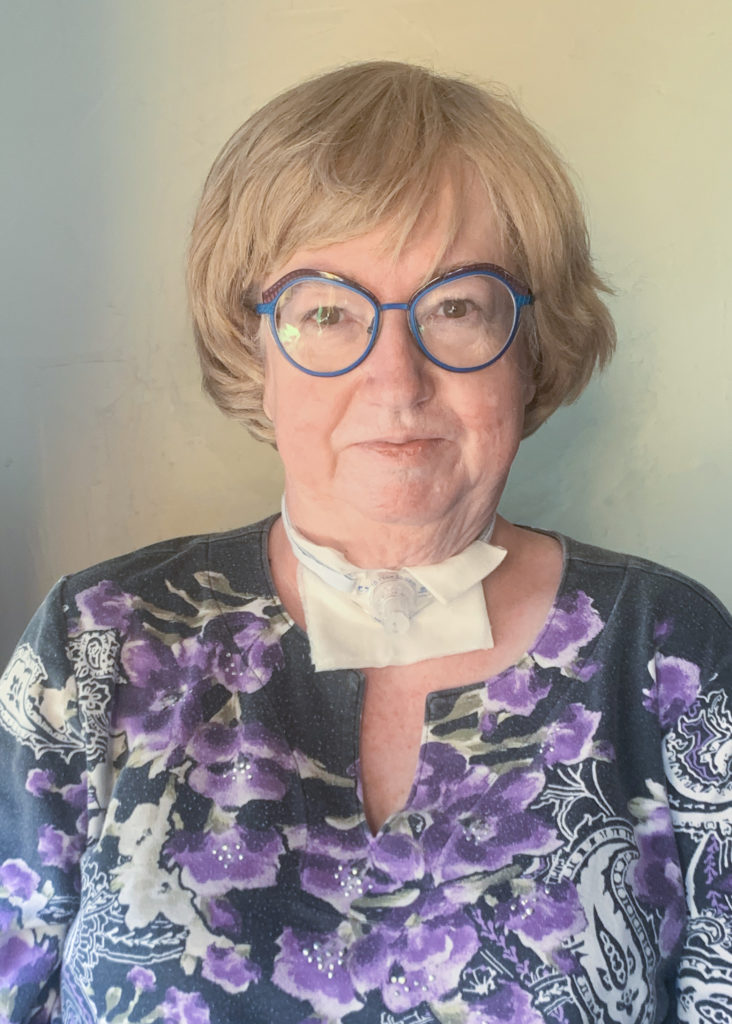 Head shot of an older woman with short blonde hair. She's earing glasses, a purple flowered dress and has a trach.