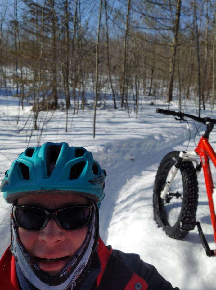 A person wearing winter gear, sunglasses and helmet, takes a selfie outside on a snowy path, with a red mountain bike in the background.