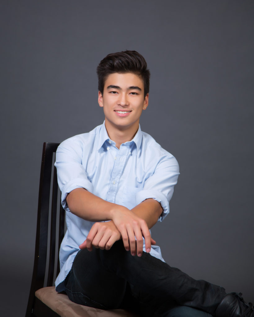 A smiling young male with dark hair sitting on a chair in front of a grey background
