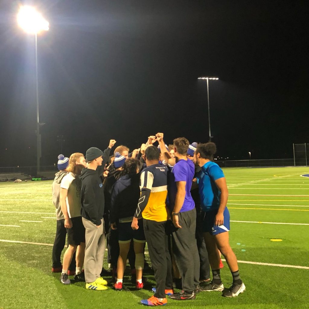 A group of people dressed casually and some in rugby uniforms, stand together on a playing field and put their hands together in the centre.
