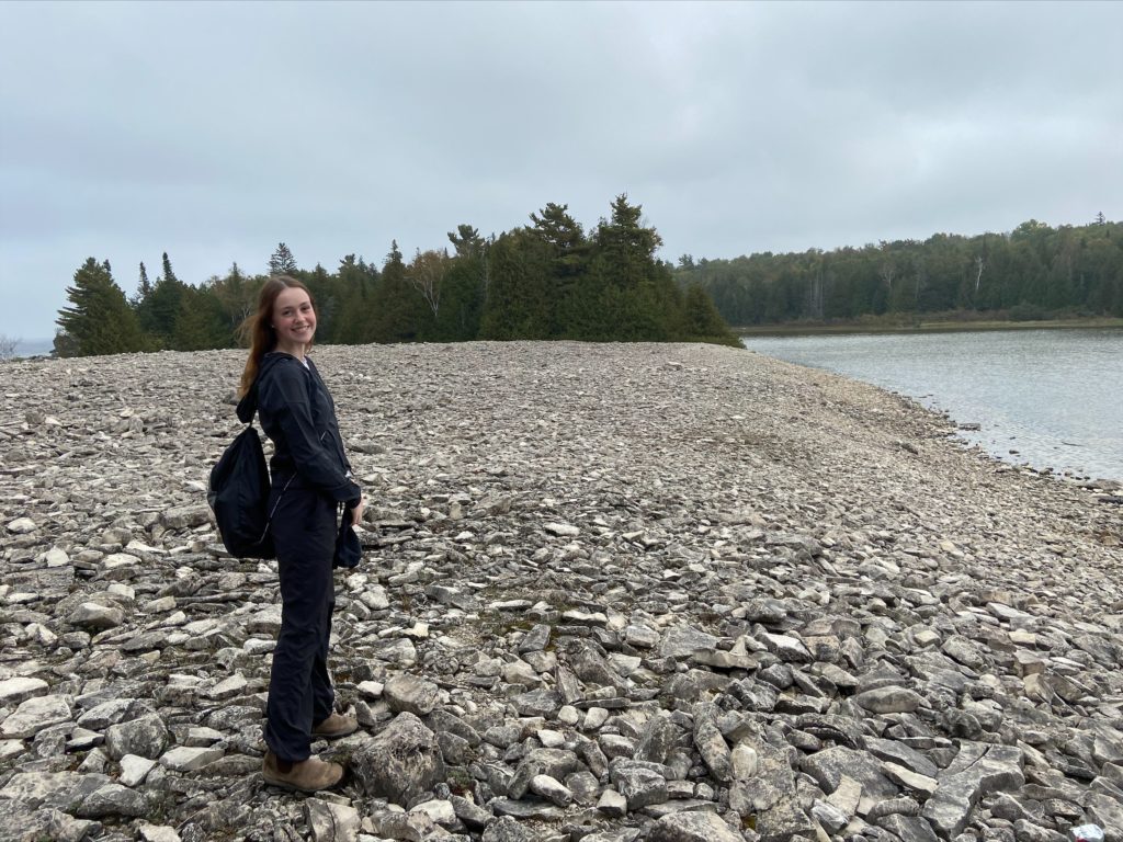 A person with long brown hair, black sweater, pants, brown shows, and backpack slung over right shoulder, stands on a rocky waterfront with green trees in the background.