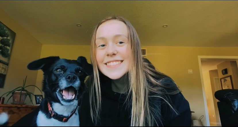 A person with long blonde hair and black sweater smiles for the camera with a small black-and-white dog.