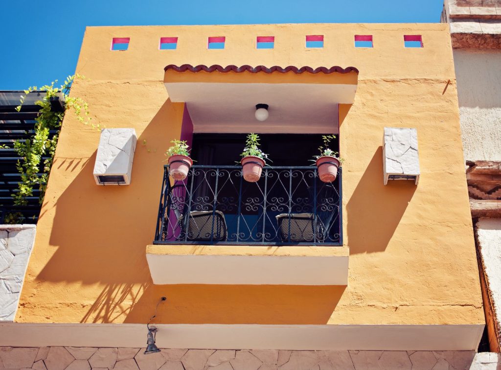 A balcony against a yellow wall, with flowerpots along the railing.
