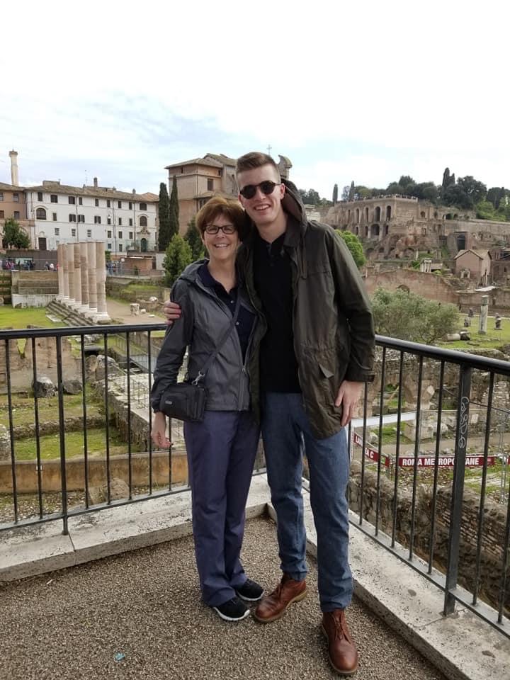 Two people stand outside on a balcony with a picturesque background of European ruins.
