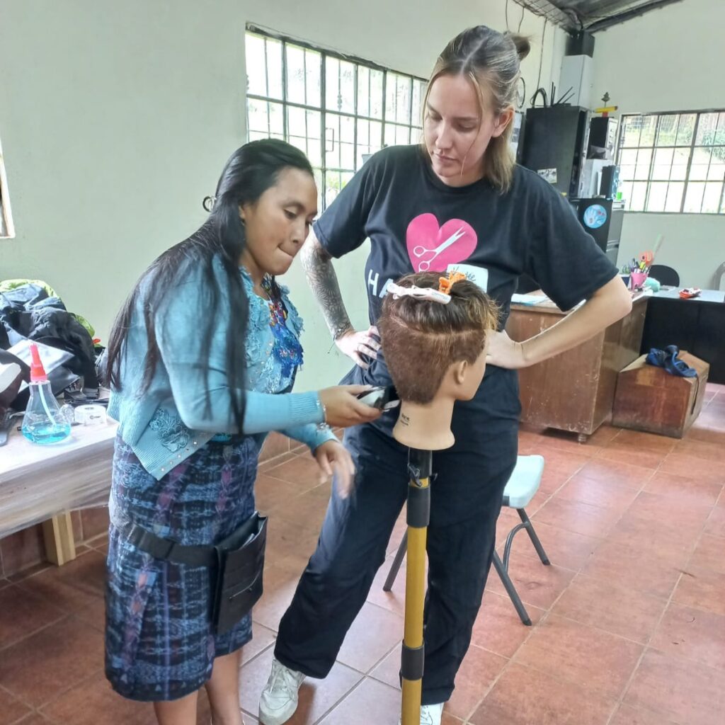 Hairstyling diploma student Kaylee Magani teaches a Guatemalan woman how to cut hair during the 2024 Hair Aid trip. Kailey has shoulder-length dark brown hair that is pulled into a half bun. She's wearing a black Hair Aid t-shirt that has a pink heart in the middle of the shirt with white scissors inside the heart. 