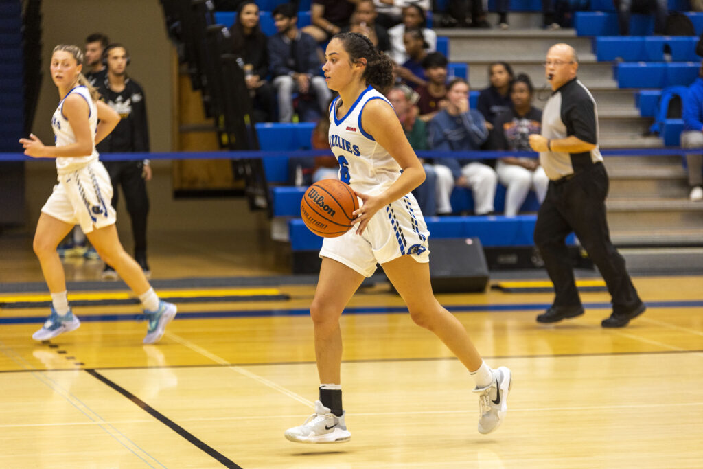 A woman with dark hair in a low bun, wearing a white and blue basketball uniform, dribbling a basketball on a court with blue bleachers in the background.