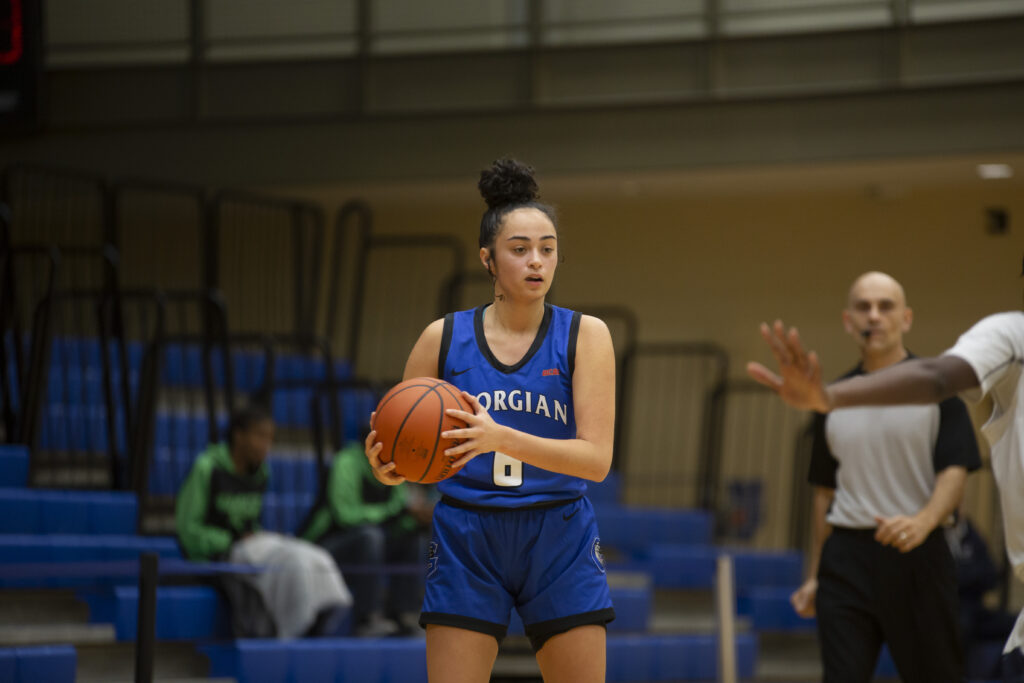 A woman with dark hair in a bun holds a basketball, wearing a dark blue Georgian Grizzlies basketball uniform, with bleachers and referee in the background.