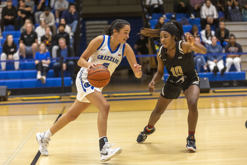 Two women on a basketball court, one dribbling the basketball wearing a blue and white uniform, the other is defending, wearing a black uniform.