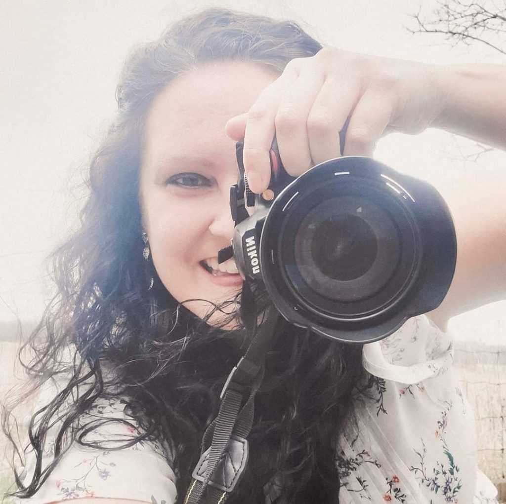 Person smiles at a camera while holding up a camera to their face as if they're about to take a photo.
