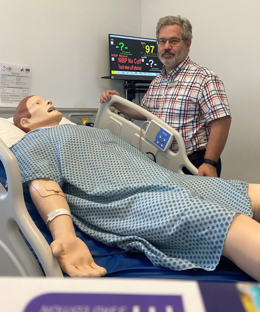 A person smiles at the camera while standing next to a patient simulator in a hospital bed.