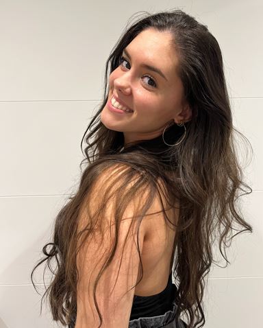 A side view of a person with long, wavy brown hair, hoop earrings, black top and jeans, turning toward the camera and smiling.