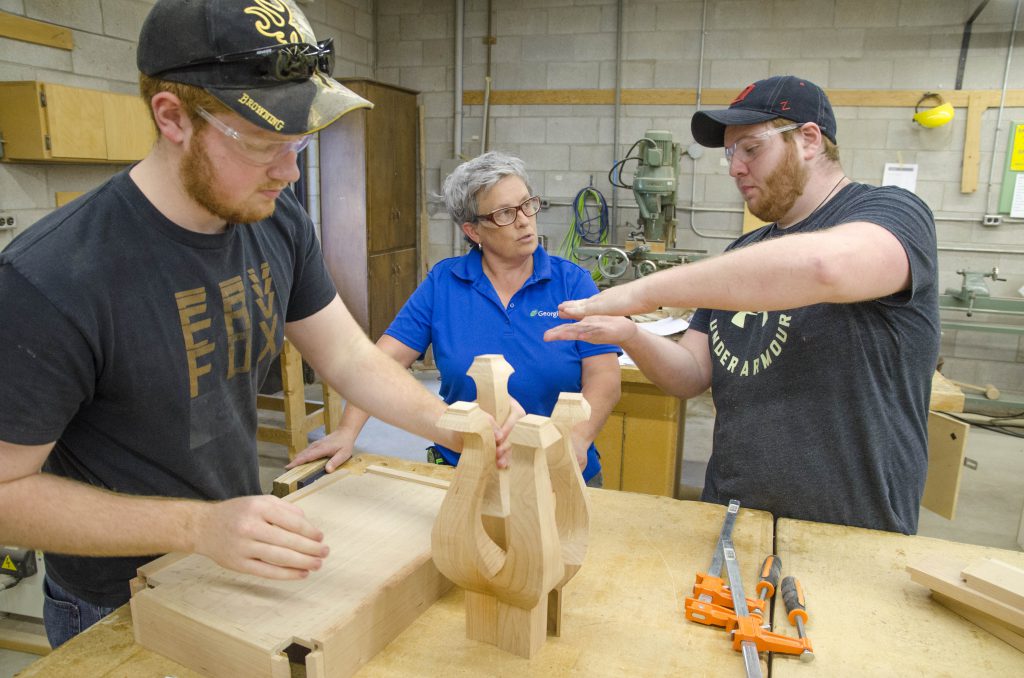 Three people stand in a woodshop, with one working on a project and the other two talking and gesturing with each other.