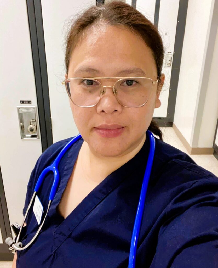 Internationally Educated Nurse Mae Jane Sanchez takes a selfie in the hallways. She's wearing her dark brown hair back in a pony tail and is wearing dark neutral scrubs with a stethoscope hanging around her neck.