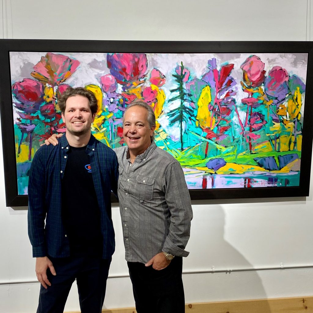 Two people dressed in black pants and collared shirts stand next to each other, arms over each other's shoulders, in front of a large colourful painting of a nature landscape.