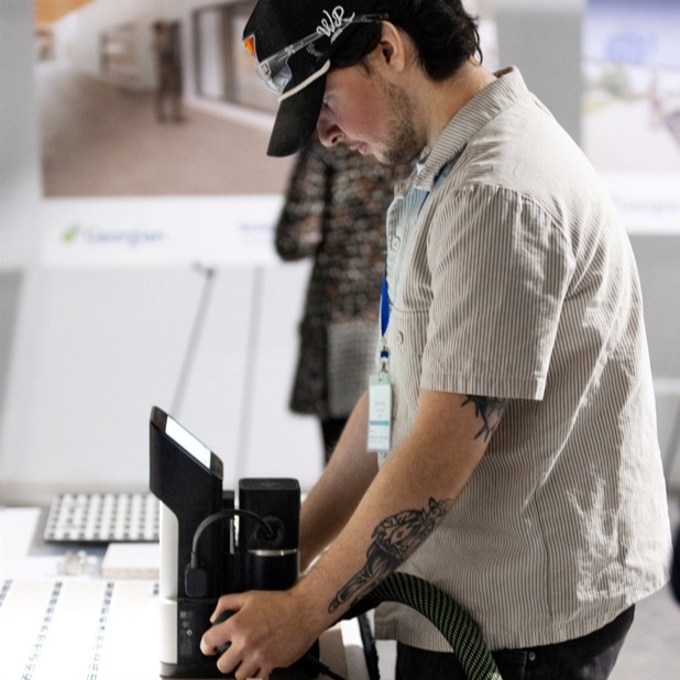 Cabinetmaking Techniques students Matthew Rees works on a project in the shop space at the Barrie Campus of Georgian College. He's wearing a black baseball cap, beige corduroy short sleeved shirt and is looking down at a piece of equipment. He has dark brown hair and a light beard.