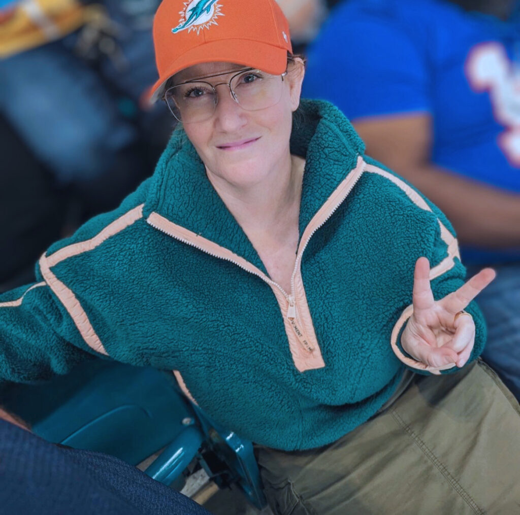 People of Georgian Melissa Smith is smiling and making a peace sign with her left hand. She's wearing an orange Miami Dolphins baseball hat, is wearing glasses and a army green cargo skirt with greet and pink-lined sweater.