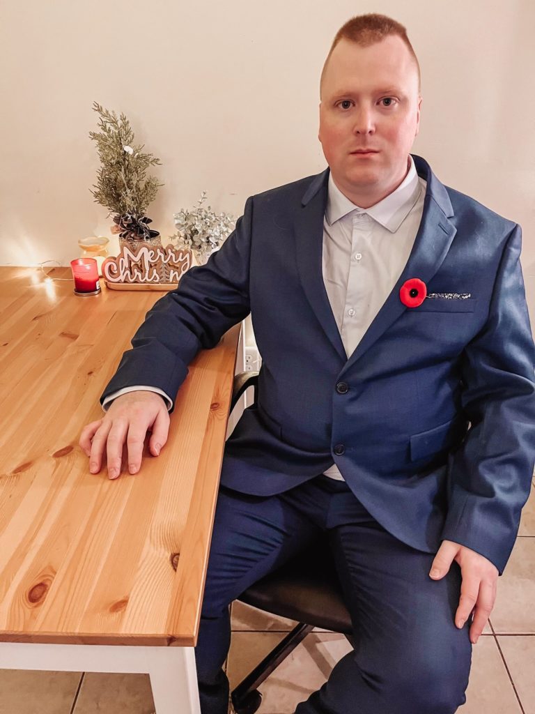 A person wearing a blue suit and a red poppy on their chest, sits at a wooden table and looks at the camera.