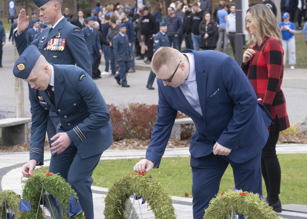 Two people, one wearing a blue military uniform, and the other in a blue suit, place poppy pins on wreaths at a ceremony.