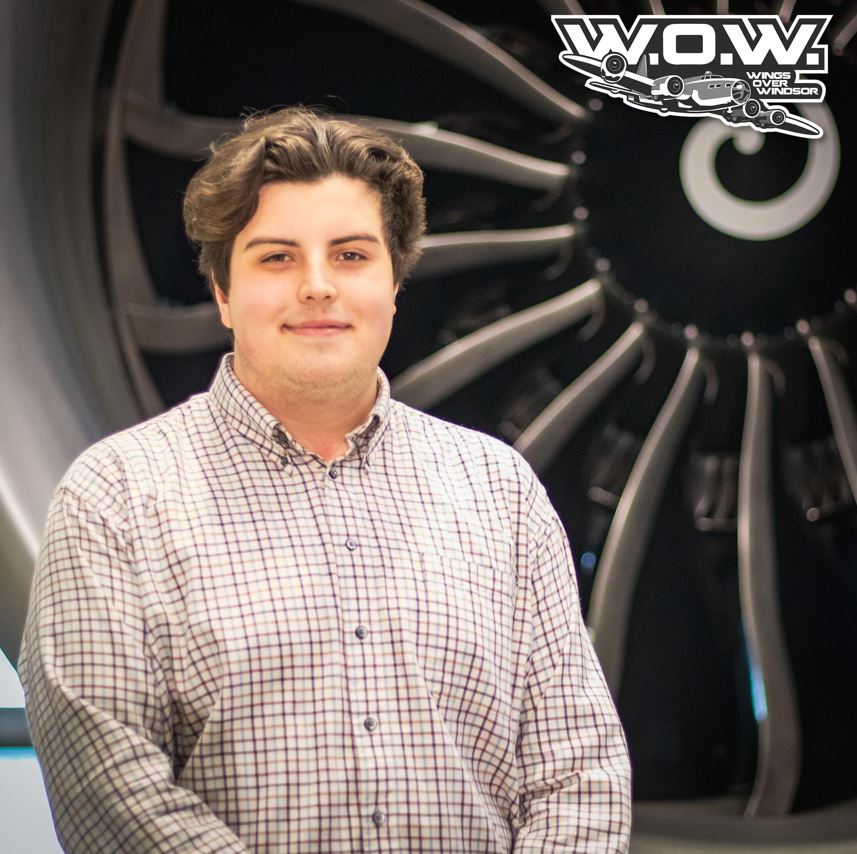 A person with brown hair, scruffy goatee, and white and red checked shirt, smiles and stands in front of an airplane engine.