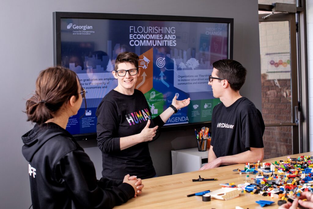 Three people interact with each other next to a table covered in LEGO pieces and a TV screen reading "Flourishing Economies and Communities."