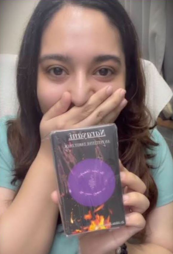 A person with long brown hair, green shirt, and their right hand over their mouth, looks at the camera and holds up a Tarot card deck with their other hand.