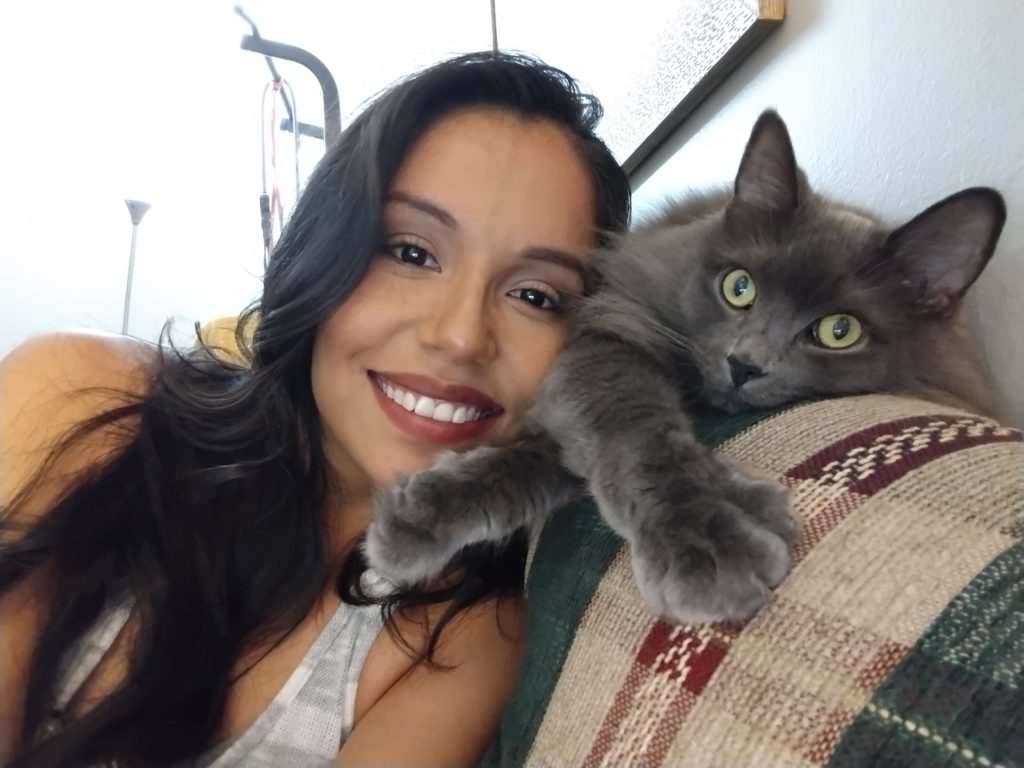 A person with long black hair and a white-and-grey tank top leans against the back of a couch with their head on near a grey cat that's lying across the top of the couch.