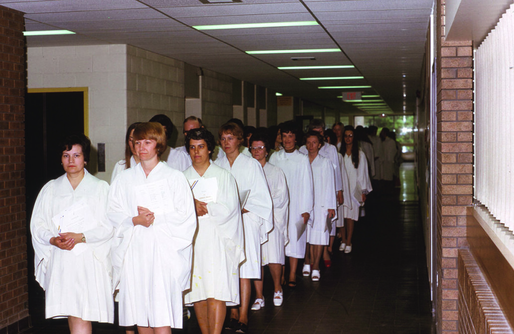 Two dozen nursing students in white gowns line up in pairs in a corridor at a convocation ceremony in 1974.