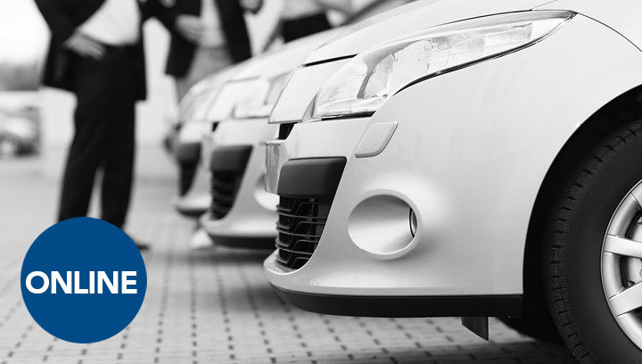 The front bumper of several automobiles lined up, with a group of people in business suits standing to the side