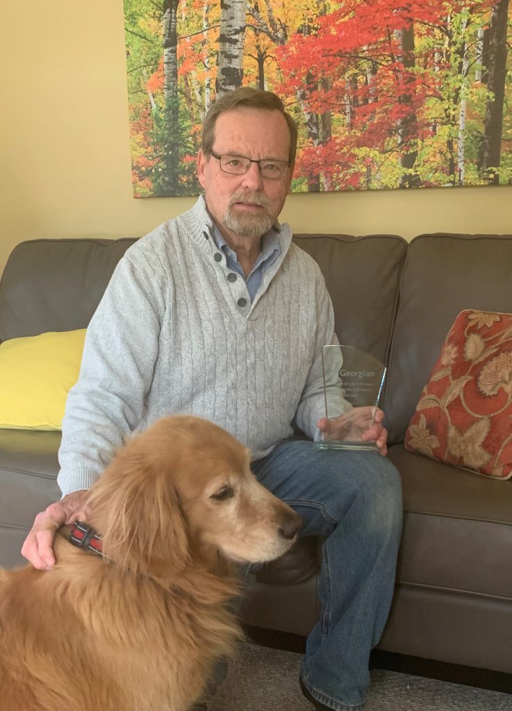 A person sits on a couch and looks at the camera, while holding an award in one hand and petting a dog with the other.