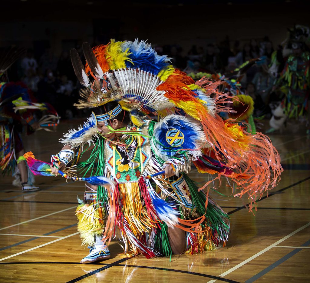 An Indigenous person dressed in traditional multicoloured headdress and garments, performing a dance. 
