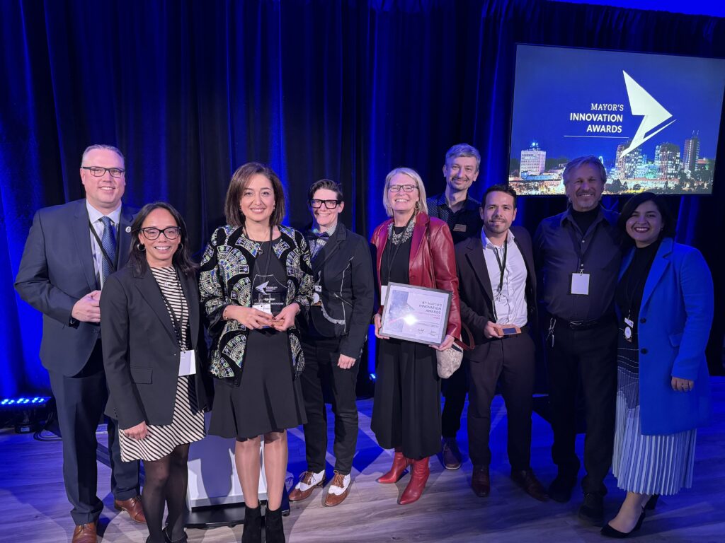 Nine people stand together in front of a screen reading "Mayor's Innovation Awards."