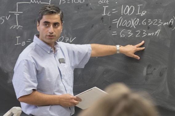 Instructor explaining electrical engineering calculations on a chalkboard to students in a classroom.